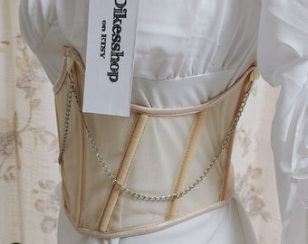 May include: A beige mesh corset with a silver chain detail. The corset is worn over a white blouse. The tag on the corset reads "Dikessshop on ETSY".