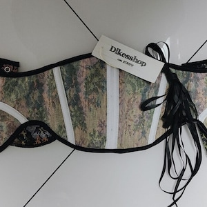 May include: A patterned corset with a tag that reads "Dikesshop on Etsy." The corset features a camouflage-like design with white accents and black trim. Black ribbons are attached to the side.