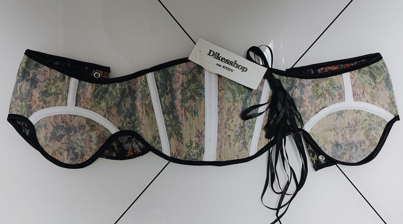 May include: A patterned corset with a tag that reads "Dikesshop on Etsy." The corset features a camouflage-like design with white accents and black trim. Black ribbons are attached to the side.