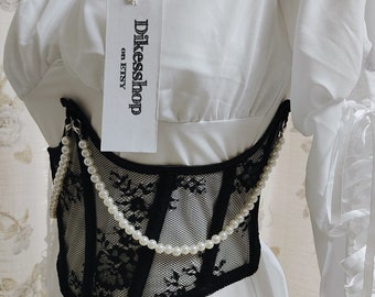 May include: Black lace corset with a pearl chain detail. The corset is worn over a white off-the-shoulder blouse. The tag on the blouse reads "Dikessshop on ETSY".