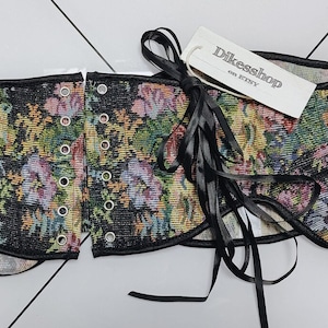 May include: A floral tapestry corset with black trim and eyelets. The corset features a colorful floral pattern with pink, yellow, and green flowers on a black background. A tag with the text "Dikesshop on Etsy" is attached with a black ribbon.
