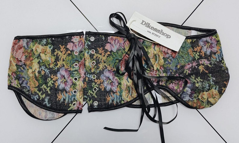 May include: A floral tapestry corset with black trim and eyelets. The corset features a colorful floral pattern with pink, yellow, and green flowers on a black background. A tag with the text "Dikesshop on Etsy" is attached with a black ribbon.