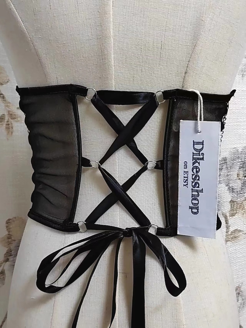 K&ouml;nnte beinhalten: Schwarzes Mesh-Korsett mit Satinband-Schn&uuml;rung und silbernen Metallringen. Das Korsett hat eine schwarze Satinband-Schleife auf der R&uuml;ckseite. Das Etikett lautet "Dikeshop on Etsy".