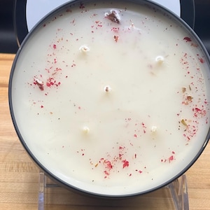 May include: A round, black candle tin with a white candle inside. The candle has six wicks and is decorated with red and brown dried flower petals. The lid has the text "Sarah Lane Candle Co" and "Five Star Resort".
