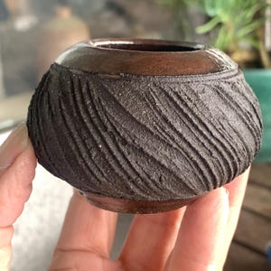 May include: A small, handcrafted ceramic pot with a dark brown, textured exterior and a smooth, darker rim. The pot's surface features a diagonal, ridged pattern. The interior is dark, and the pot is held in a hand.