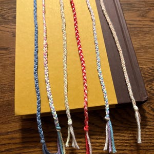May include: Seven colorful braided cords with knotted ends. The cords are made of different colors of yarn, including blue, pink, yellow, white, and red.