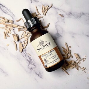 May include: A brown glass bottle with a dropper top containing Ashwagandha Tincture from Hillside Botanicals. The bottle label is cream colored with brown text and a graphic of a plant. The text on the label reads "ESTD 2018 HILLSIDE BOTANICALS ASHWAGANDHA TINCTURE stress & vitality support HERBAL SUPPLEMENT 1 OZ/30ML".