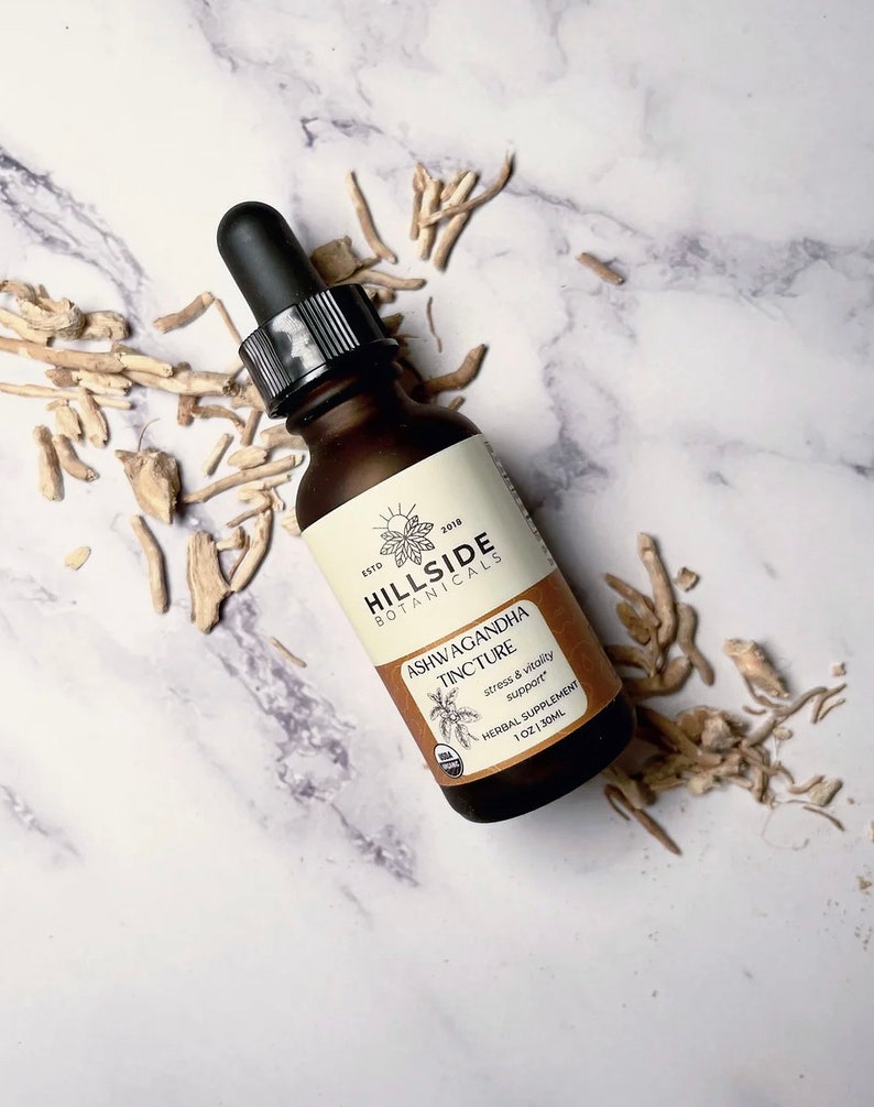 May include: A brown glass bottle with a dropper top containing Ashwagandha Tincture from Hillside Botanicals. The bottle label is cream colored with brown text and a graphic of a plant. The text on the label reads "ESTD 2018 HILLSIDE BOTANICALS ASHWAGANDHA TINCTURE stress & vitality support HERBAL SUPPLEMENT 1 OZ/30ML".