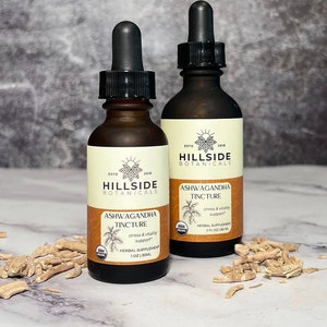 May include: Two brown glass bottles with droppers, labeled "Hillside Botanicals Ashwagandha Tincture". The bottles are on a white surface with dried root pieces scattered around them. The labels state "stress & vitality support" and include the USDA Organic seal.