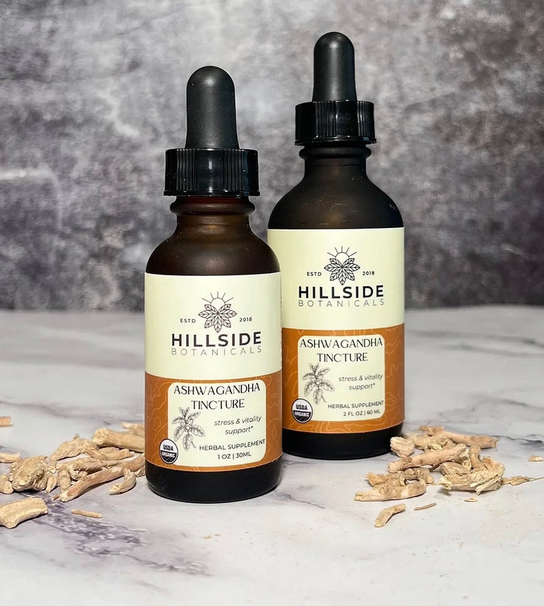 May include: Two brown glass bottles with droppers, labeled "Hillside Botanicals Ashwagandha Tincture". The bottles are on a white surface with dried root pieces scattered around them. The labels state "stress & vitality support" and include the USDA Organic seal.