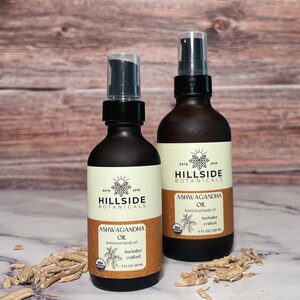 May include: Two amber glass bottles with black pump tops and labels. The labels are white with black text and a green logo. The text on the labels reads "Hillside Botanicals" and "Ashwagandha Oil". The bottles are sitting on a brown wooden surface with dried roots scattered around them.