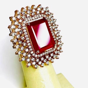 May include: A rose gold ring featuring a large, rectangular, deep red gemstone at the center. The gemstone is surrounded by a halo of small, clear, round gemstones. The ring is displayed on a light yellow stand.