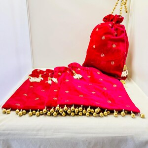 May include: A collection of three red velvet drawstring bags with gold embellishments. The bags feature gold floral embroidery and are adorned with gold beaded tassels. One bag has a gold chain handle.