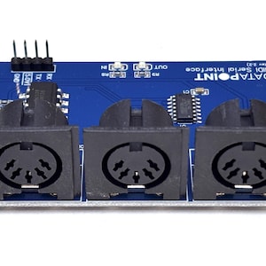 Op de afbeelding: Een blauwe rechthoekige printplaat met het label DATAPOINT MIDI Serial Interface (Rev 2.2), voorzien van drie zwarte vijfpolige DIN-connectoren op een rij, een vierpolige header en diverse geïntegreerde schakelingen op het oppervlak.