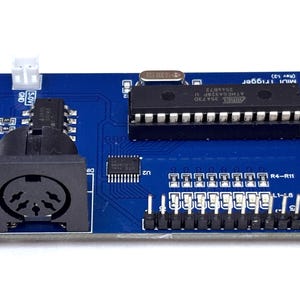 Op de afbeelding: Een blauwe printplaat met een zwarte MIDI-poort, een grote geïntegreerde schakeling, een kleinere opbouwchip, een witte connector en een rij metalen pinnen. De printplaat is voorzien van witte labels, waaronder MIDI Trigger en 5.0V.