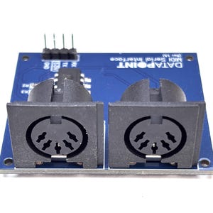 Puede incluir: Una placa de circuito impreso azul con dos puertos MIDI negros de cinco pines montados en la parte frontal. La placa está etiquetada como DATAPOINT MIDI Serial Interface (Rev 3b) e incluye un conector de cuatro pines en el borde superior.