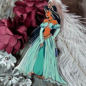 May include: A teal blue and gold enamel pin featuring a cartoon depiction of Princess Jasmine from Disney's Aladdin. She is wearing a blue and gold outfit and a jeweled headpiece.