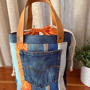 May include: A bucket bag constructed from patchwork denim and orange patterned fabric, featuring a central denim pocket, tan leather handles secured with metal rivets, and a white rope drawstring closure that cinches the orange fabric top.