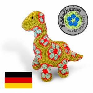 Op de afbeelding: Een groen gehaakt dinosaurus speelgoed met rode en blauwe bloemenaccenten. Het speelgoed staat op een witte achtergrond. De afbeelding bevat ook een Duitse vlag en een rond zilveren label met de tekst "Häkelhaken aus Leidenschaft".