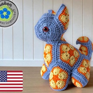 Könnte beinhalten: Eine handgefertigte blaue Häkelkatze mit orangefarbenen und gelben Akzenten. Die Katze hat große rote Augen und ist mit einem Granny-Square-Muster gefertigt. Das Bild enthält auch ein Logo mit deutschem Text und eine US-Flagge.