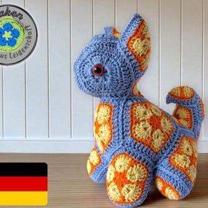 Könnte beinhalten: Handgehäkelte Katze in Blau, Orange und Gelb. Die Katze hat große rote Augen und einen Schwanz. Das Bild enthält eine deutsche Flagge und ein Logo mit Text. Die Katze steht auf einer Holzoberfläche.