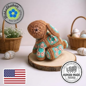 Op de afbeelding: Een handgemaakt gehaakt konijn in bruin, roze en teal, met een bloemmotief, zittend op een houten schijf. De afbeelding bevat een mand met eieren en een sticker met de tekst "100% Met zorg gemaakt".