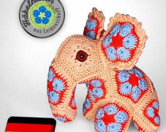 Op de afbeelding: Een gehaakt olifantenspeelgoed met een roze lichaam en blauwe en rode bloemenaccenten. De olifant heeft een zwart oog en zit op een wit oppervlak. De afbeelding bevat ook een Duitse vlag en een rond metalen label met de tekst "Häkelhaken aus Leidenschaft".