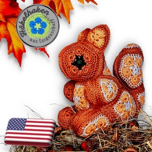 Könnte beinhalten: Ein gehäkeltes Eichhörnchenspielzeug mit orangefarbenen und weißen Akzenten, das in einem Heunest sitzt. Das Eichhörnchen hat ein schwarzes Knopfauge und ist von Herbstblättern und Eicheln umgeben. Das Bild zeigt auch eine kleine amerikanische Flagge.