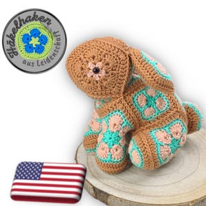 Könnte beinhalten: Ein handgehäkelter Hase in Braun-, Türkis- und Rosatönen. Der Hase hat eine Blume auf dem Kopf und dekorative Quadrate auf dem Körper. Der Hase sitzt auf einer Holzoberfläche.