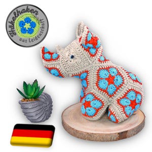 May include: A crocheted rhinoceros toy with blue and orange flower patterns. The toy is sitting on a wooden base. The background includes a small potted plant and a German flag.