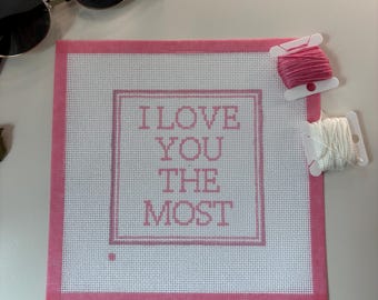 May include: A square, pink-bordered canvas with the text "I LOVE YOU THE MOST" in pink. Two spools of thread, one pink and one white, are next to the canvas. A pair of sunglasses with gold frames and black lenses is also in the image.