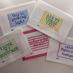 May include: Five rectangular needlepoint canvases featuring travel-themed phrases. The designs include blue stripes with 'this is packing light', yellow stripes with 'ARE WE THERE YET?', green stripes with 'I LOVE MY SUITCASE', pink stripes with 'where to next?', and a purple border with 'catch flights,not feelings'.