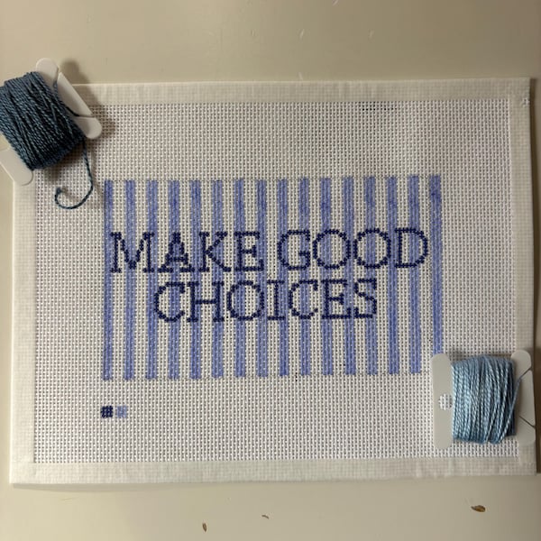 Può includere: Una tela rettangolare bianca per ricamo con strisce verticali azzurre e il testo blu scuro "MAKE GOOD CHOICES" ricamato al centro. Due bobine di plastica con filo da ricamo blu sono posizionate in alto a sinistra e in basso a destra.