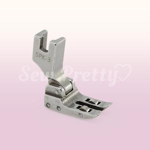 Puede incluir: Un pie de máquina de coser de plata con una abertura en forma de U y el texto "SPK-3" grabado en el lateral.