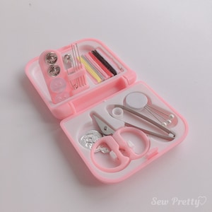 Pode incluir: Um kit de costura de viagem rosa com uma variedade de ferramentas, incluindo tesoura, agulhas, linha, botões e um almofada para alfinetes. O kit está aberto e as ferramentas são visíveis.