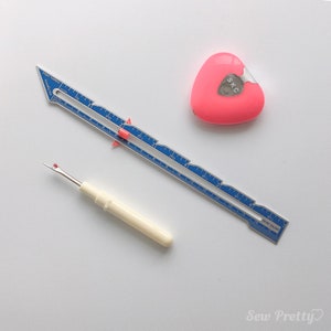 May include: A blue seam gauge ruler with a red marker, a white seam ripper with a red tip, and a pink heart-shaped fabric marker with the text "SKC" on it.