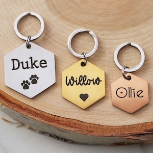 May include: Three hexagonal dog tags with key rings. The tags are engraved with the names "Duke", "Willow", and "Ollie". The Duke tag is silver, the Willow tag is gold, and the Ollie tag is copper.