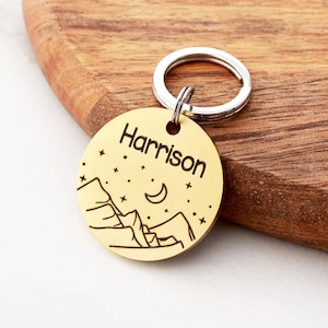 May include: Gold-colored metal dog tag with a mountain and moon design and the name "Harrison" engraved on it.