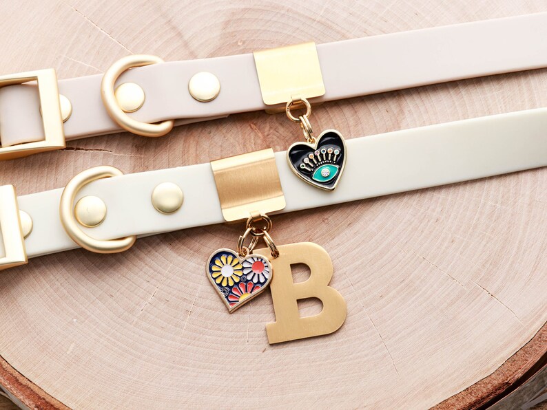 May include: Two white leather dog collars with gold hardware and charms. The top collar has a black heart charm with an evil eye design. The bottom collar has a gold letter B charm and a heart charm with a floral design.
