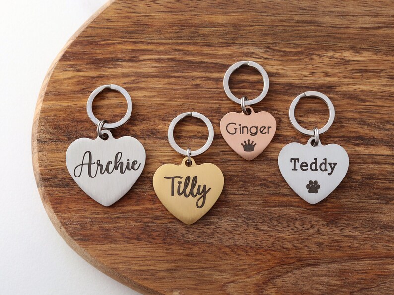 May include: Four heart-shaped dog tag keychains with engraved names: Archie, Tilly, Ginger, and Teddy. The keychains are silver, gold, rose gold, and silver with a paw print.