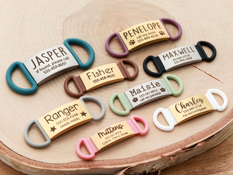 May include: A collection of personalized pet ID tags in various colors, including teal, brown, and pink. Each tag features a name and contact information, with some including additional details like "needs meds."