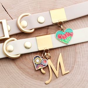 May include: Two white leather dog collars with gold hardware and charms. One charm is a heart with red tulips on a green background. The other charm is a heart with colorful flowers and the letter M.