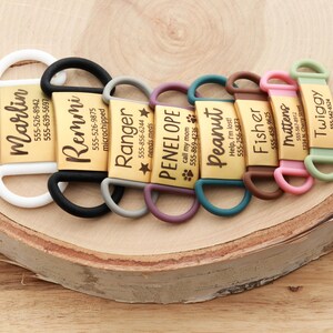 May include: A collection of personalized pet ID tags with names and phone numbers. Each tag is a gold rectangle with a different name in black script and a colored silicone loop. The tags are arranged on a wooden surface.