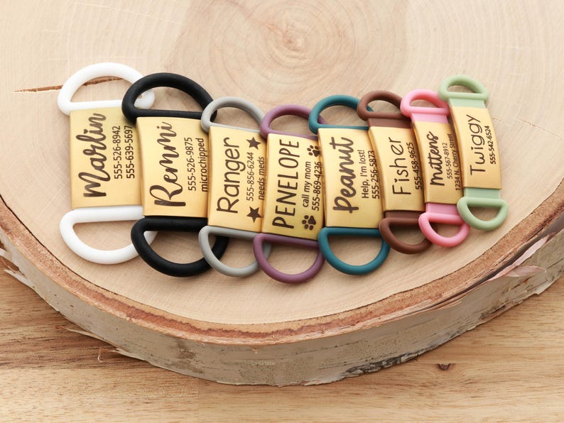 May include: A collection of personalized pet ID tags with names and phone numbers. Each tag is a gold rectangle with a different name in black script and a colored silicone loop. The tags are arranged on a wooden surface.