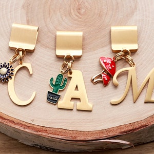 May include: Three gold bookmark clips with charms. The charms are a blue flower, a green cactus with the word "hello" on the pot, and a red mushroom. The clips are labeled with the letters C, A, and M.