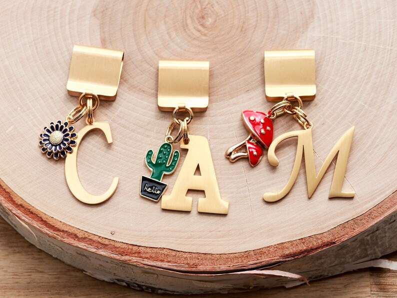 May include: Three gold bookmark clips with charms. The charms are a blue flower, a green cactus with the word "hello" on the pot, and a red mushroom. The clips are labeled with the letters C, A, and M.
