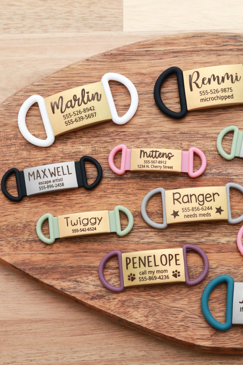 May include: A collection of personalized pet ID tags in various colors, including white, black, pink, green, and purple. Each tag features a name and contact information, with some including additional details like "microchipped" or "needs meds."