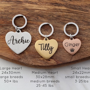 May include: Three heart-shaped dog tags with key rings. The tags are engraved with the names "Archie", "Tilly", and "Ginger". The tags are silver, gold, and rose gold, respectively. The tags are shown on a wooden surface with text describing the size and weight of each tag.