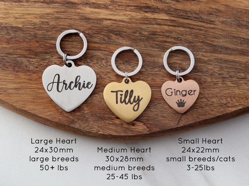 May include: Three heart-shaped dog tags with key rings. The tags are engraved with the names "Archie", "Tilly", and "Ginger". The tags are silver, gold, and rose gold, respectively. The tags are shown on a wooden surface with text describing the size and weight of each tag.