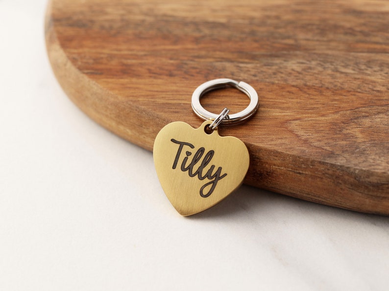 May include: Gold heart-shaped dog tag with the name "Tilly" engraved on it. The tag is attached to a silver key ring.